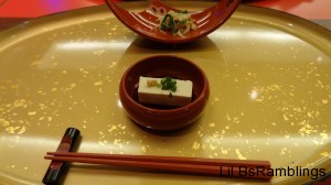 A rectangle of tofu in a bowl with a sprig of herbs next to a small salad