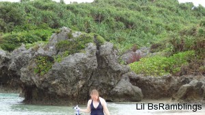 Me leaving the water with my snorkeling gear in my hands and a rock formation in front of green slope behind me.
