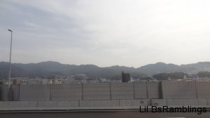 Kure as seen from over a highway fence with mountains behind under a cloudy sky.