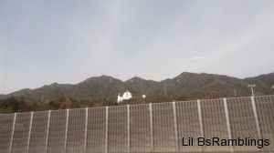 A Buddhist Shrine seen over a highway fence with tiny golden Budhas on the 3 roof points.