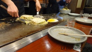 The egg cooking behind a finished stack of egg under veges, meat, and pancake.