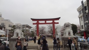 A large tori-i gate protected by a huge stone lion on each side.  Through the gate is a walkway lined with cherry trees in bloom.