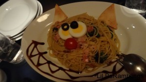 A plate of yellow noodles with bread for ears, eggs and olives for eyes, a cherry tomato for a nose, and sauce on the side forming a Cheshire Cat grin.