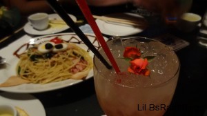 A light pink drink with tiny roses resting on the ice next to a red straw and a black staw.