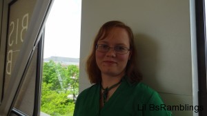 Red-haired me in a green dress and green dragonfly necklace with trees in a window behind me.