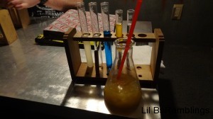 A medium sized beaker with orange juice in front of five small beakers with different colors in them on a wooden rack.