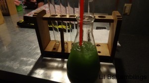 A greenish drink in a beakerin front of the wooden rack of empty smaller beakers.