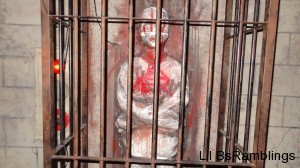 A plastic mummy highlighted with blood behind aged bars.